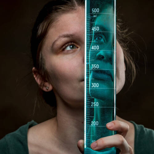 portrait of GVSU student scholar Lilly Helsel looking through a glass beaker with an effect of having her face reflected in the beaker